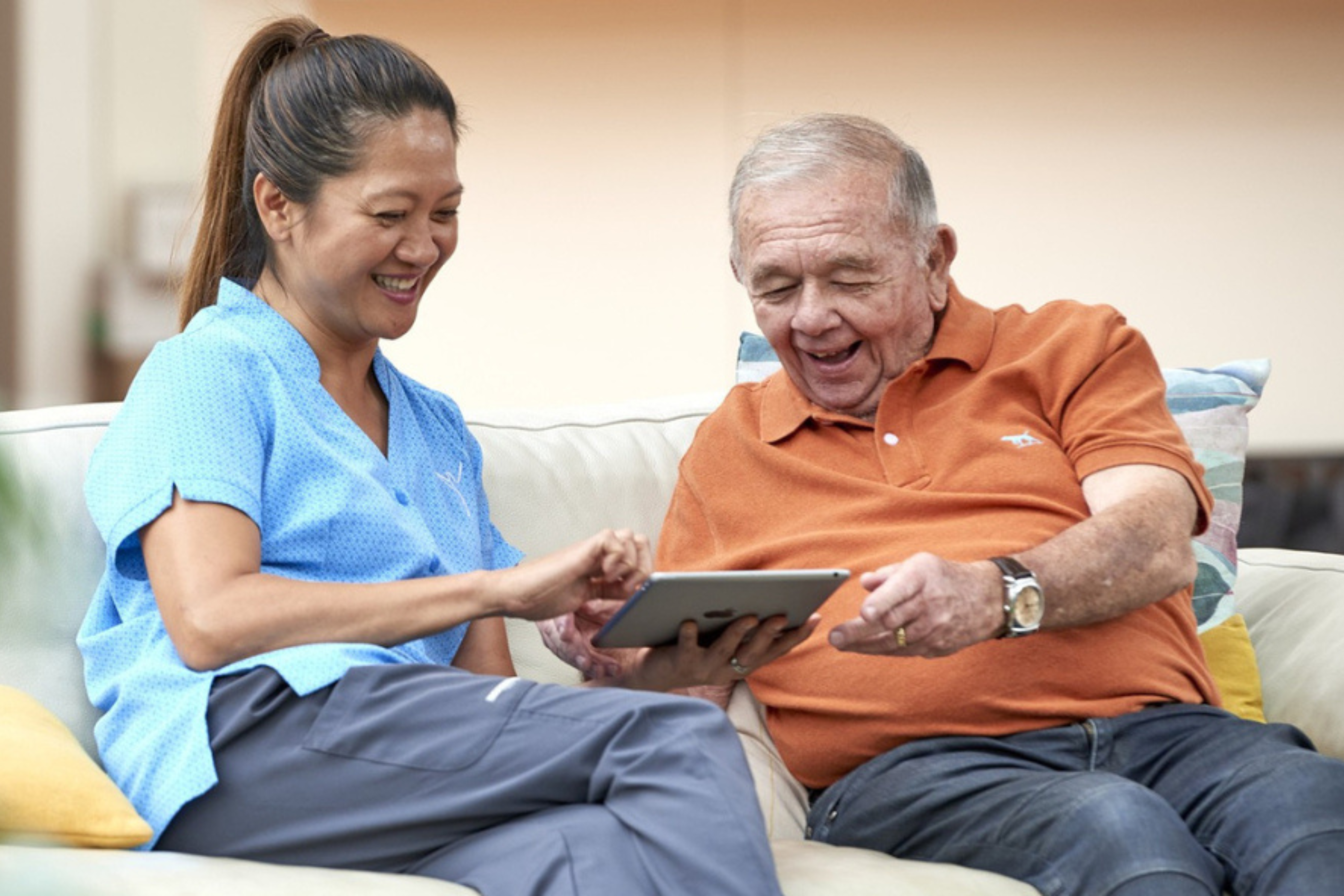 A cheerful caregiver in scrubs sits beside an older man on a couch, both engaged and smiling while interacting with a tablet. The scene highlights companionship, digital engagement, and supportive aged care in a comfortable, home-like setting.