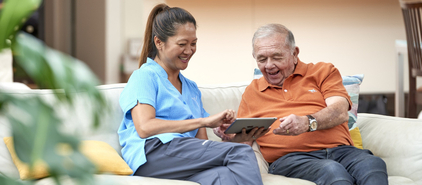 A smiling aged care worker sitting on a couch with an elderly man, both looking at a tablet together in a home-like setting.
